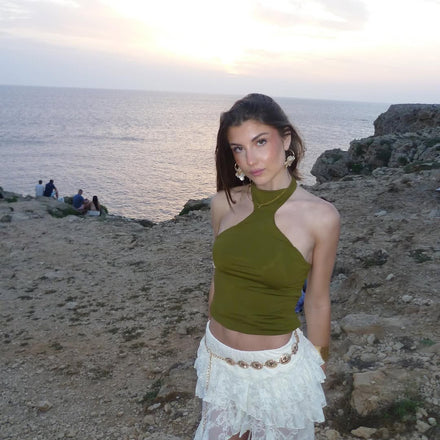 Woman in a green top and white skirt standing on a rocky beach with people in the background.